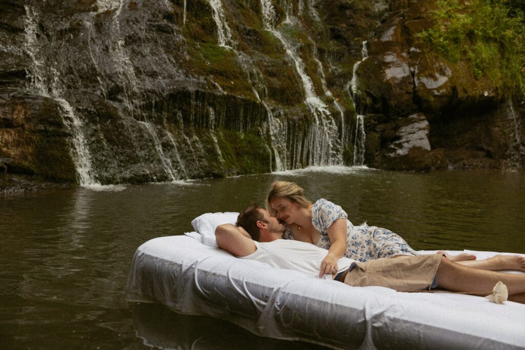 Man and woman laying on air mattress in water in front of waterfall about to kiss.