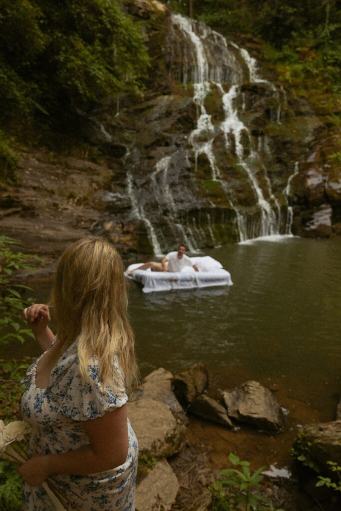 Woman looking at man while he is on air mattress in water in front of waterfall.