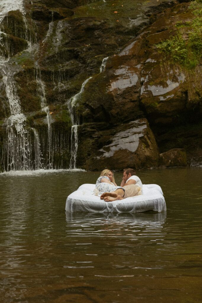 Man and woman laying and looking at each other on air mattress in water in front of waterfall.
