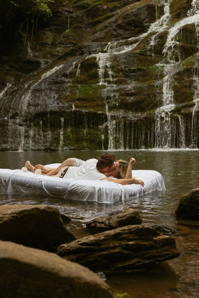 Man and woman laying on air mattress in water in front of waterfall kissing.