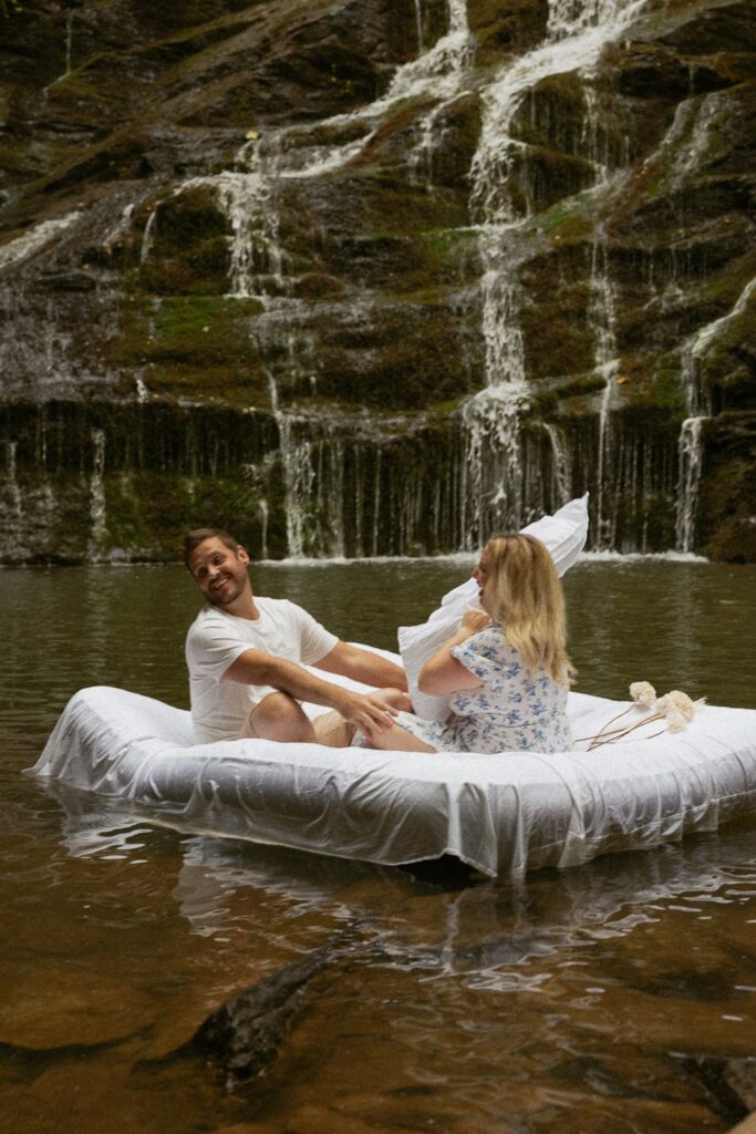 Woman hitting man with pillow while they are sitting on air mattress in water in front of waterfall.