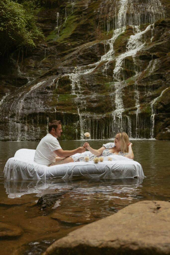 Man handing woman a flower while sitting on air mattress in water in front of waterfall.