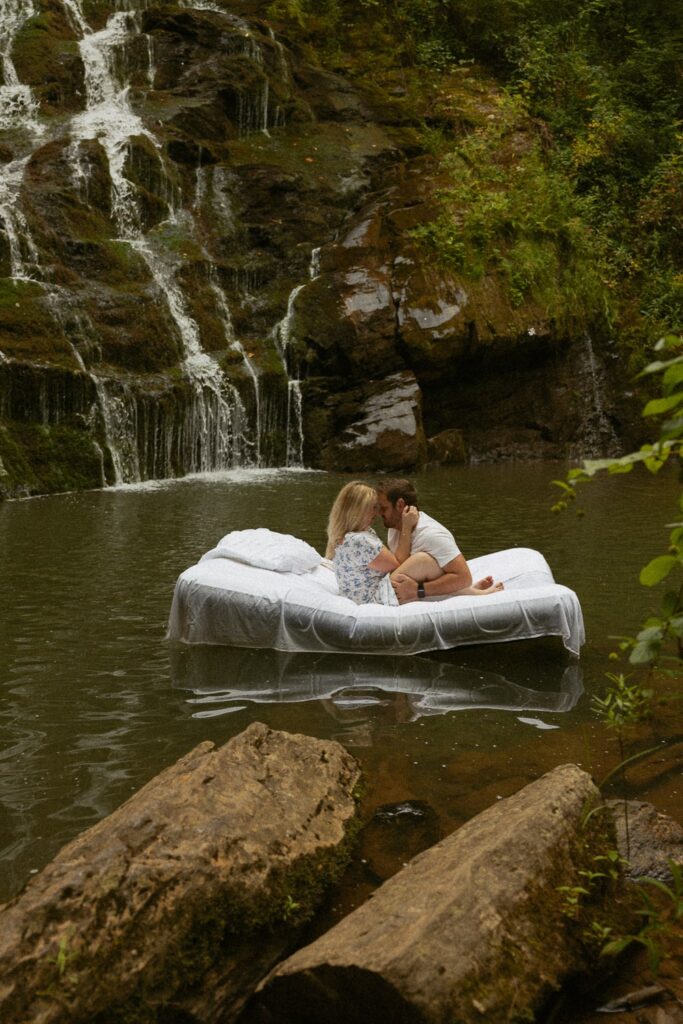 Man and woman sitting on air mattress in water in front of waterfall about to kiss.