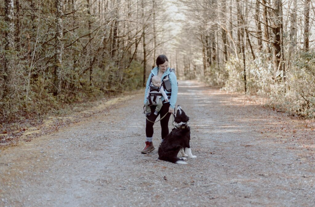 Woman standing in road with baby in baby carrier and dog on a leash.