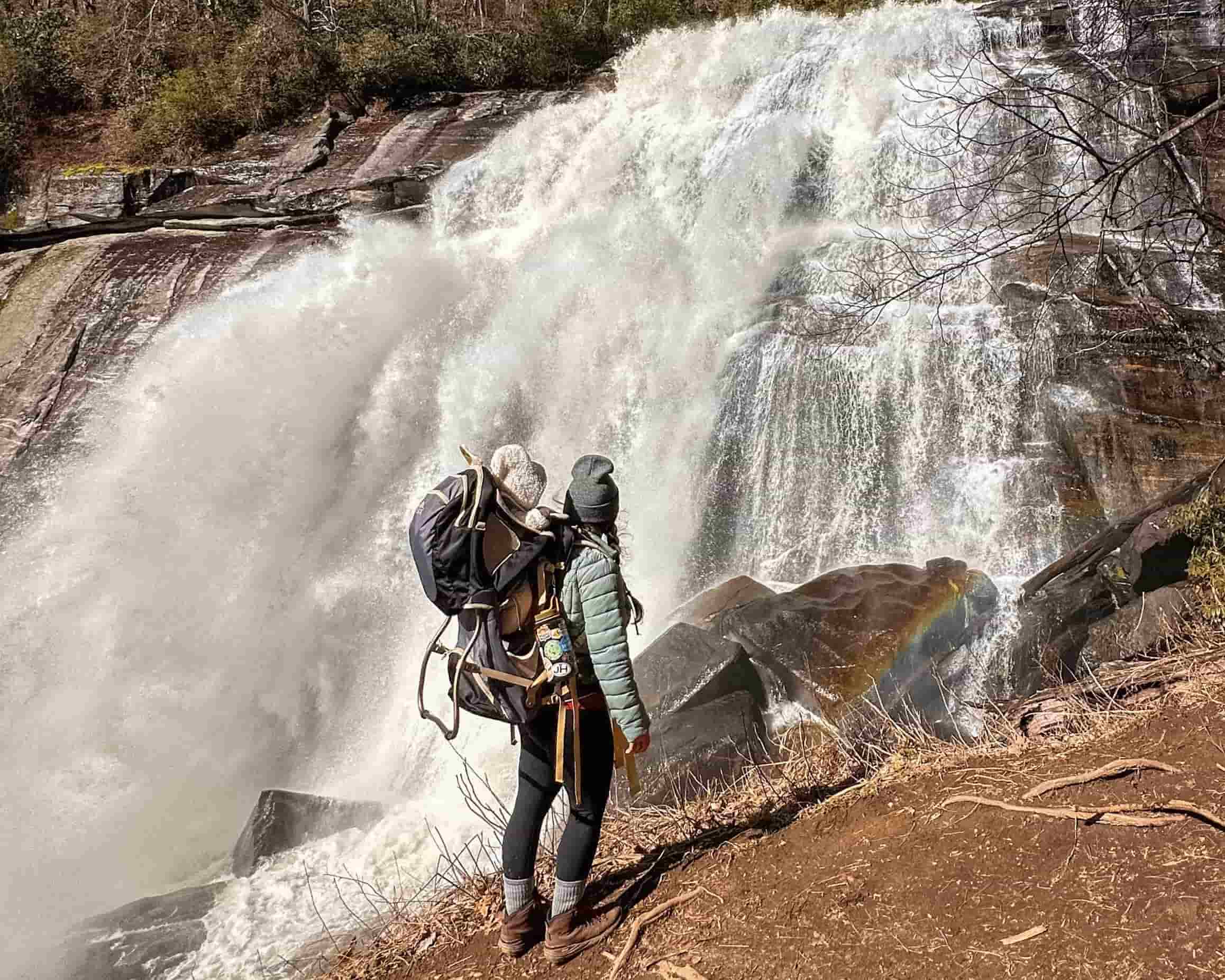 Woman with baby in hiking carrier standing in front of Rainbow Falls in North Carolina.