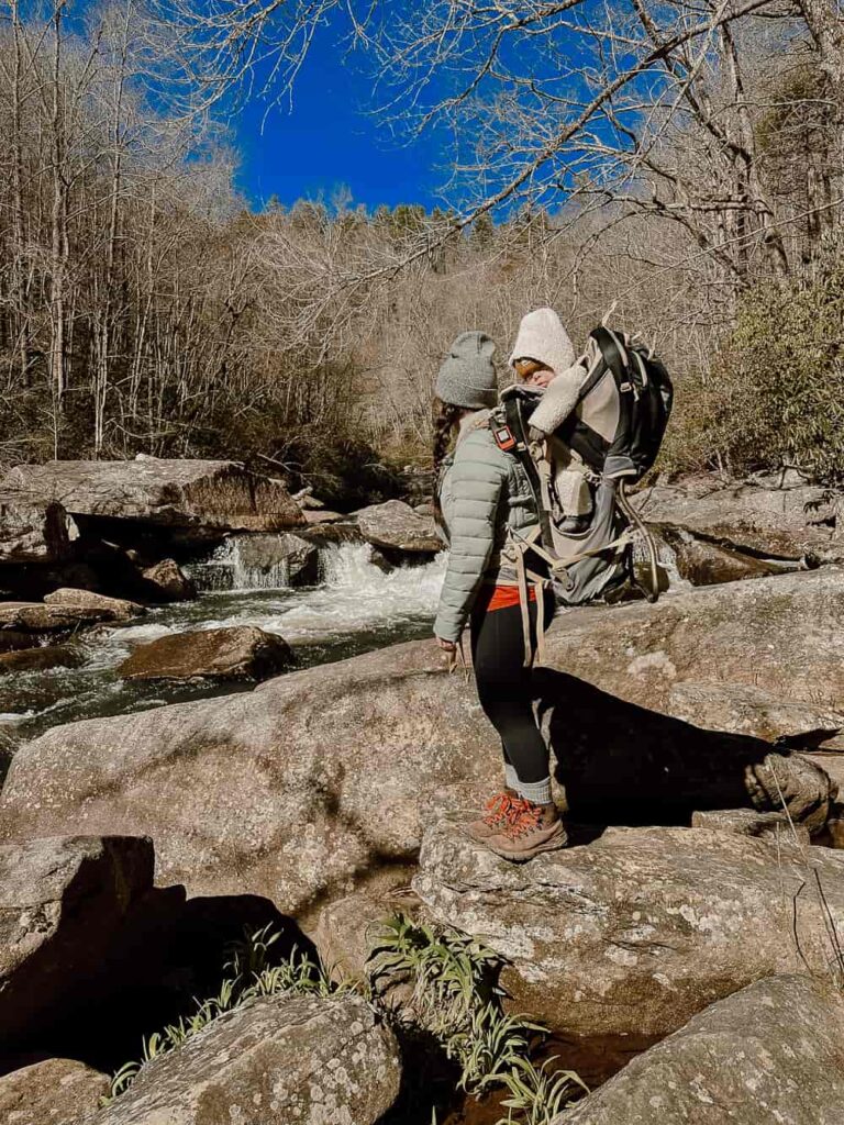Woman with baby in hiking carrier standing on rock in front of river in North Carolina.