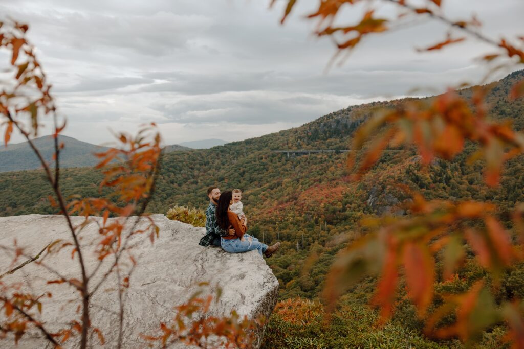 Man, woman and baby sitting on rock in front of fall colored trees at Rough Ridge overlook in Boone, NC.