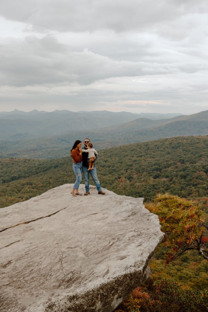 Man, woman, and baby standing on rock in front of fall colored trees at Rough Ridge overlook in Boone, NC.