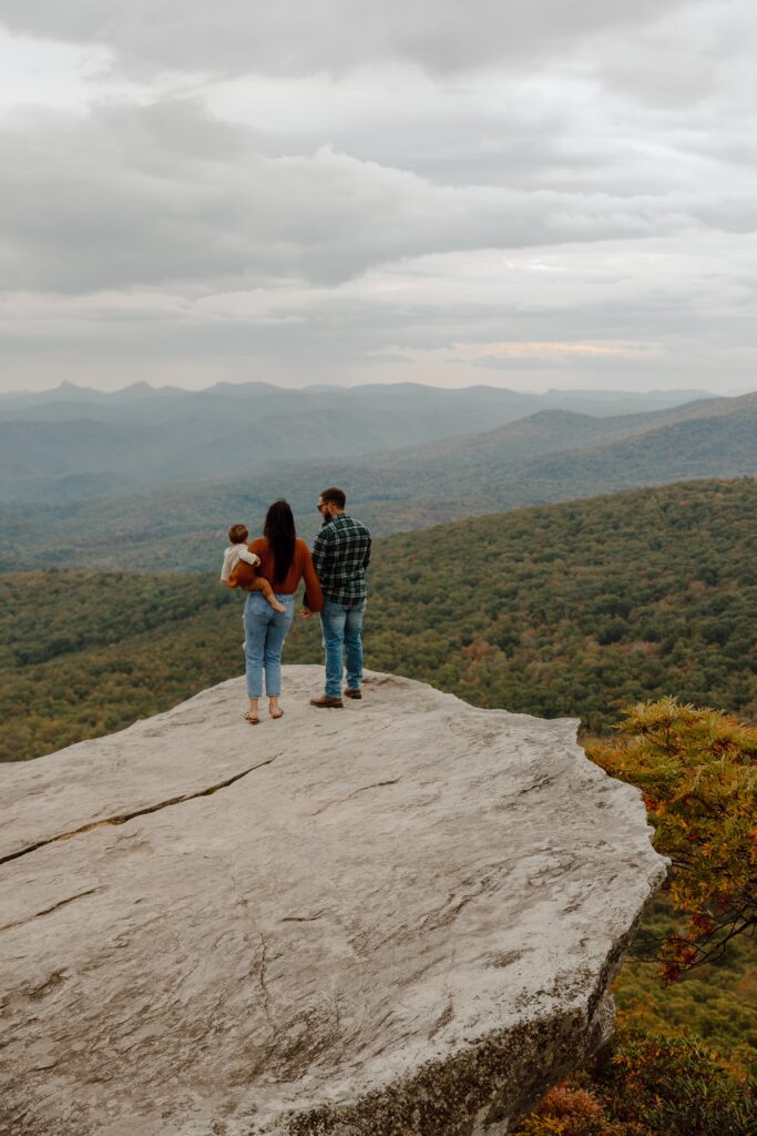Man, woman, and baby standing on rock in front of fall colored trees at Rough Ridge overlook in Boone, NC.