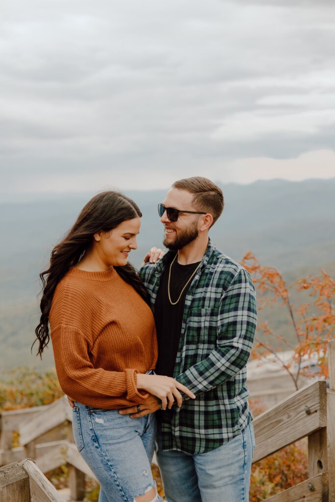 Man and woman smiling and hugging in front of fall colored trees at Rough Ridge overlook in Boone, NC.