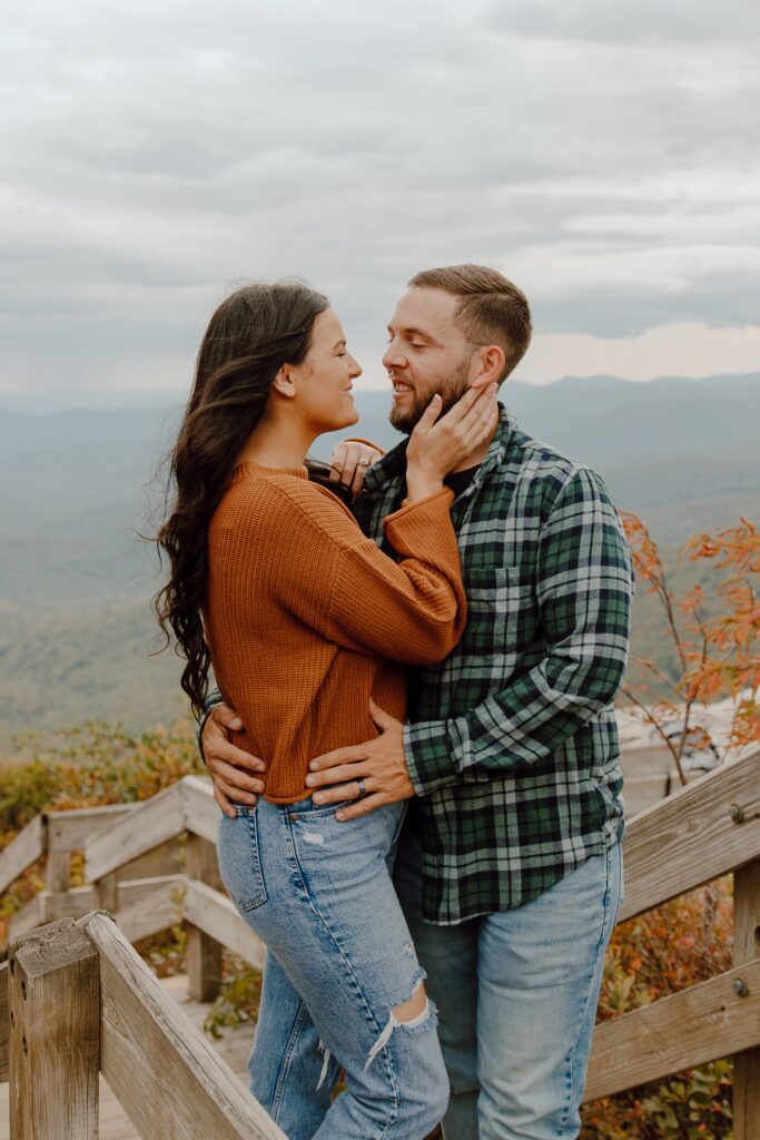 Man and woman smiling and hugging in front of fall colored trees at Rough Ridge overlook in Boone, NC.