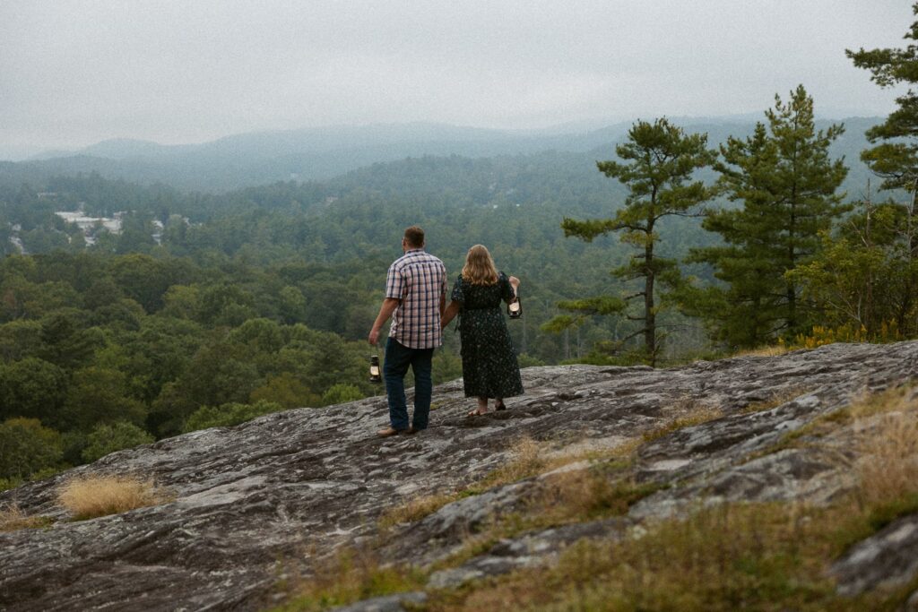 Man and woman standing on rock and holding lanterns while looking at the mountains.