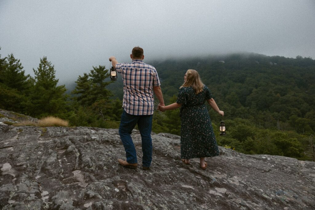 Man and woman standing on rock while holding lanterns during engagement photos.