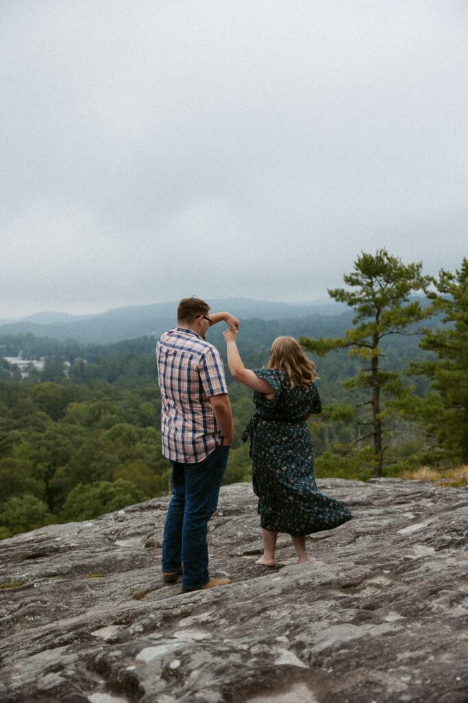 Man spinning woman on rock during engagement photos at Sunset Rock in Highlands, North Carolina.