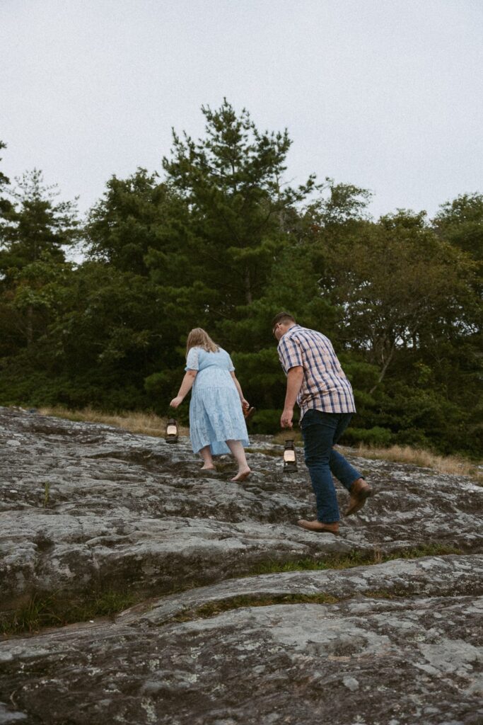 Man and woman walking up a rock while holding lanterns during engagement photos.
