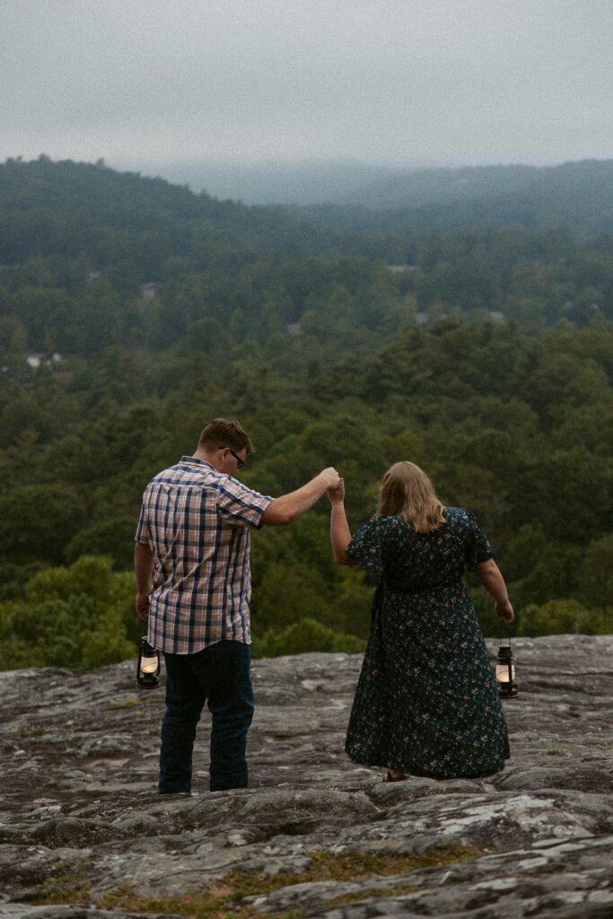 Man helping woman down rock while they hold lanterns during engagement photos at Sunset Rock in Highlands, North Carolina.