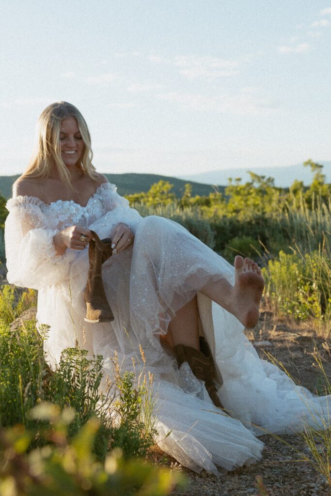 Woman smiling and putting boot on dirty feet while wearing a wedding dress.