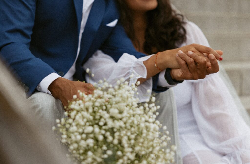 Close up photo of man and woman in wedding attire holding hands with flowers. 