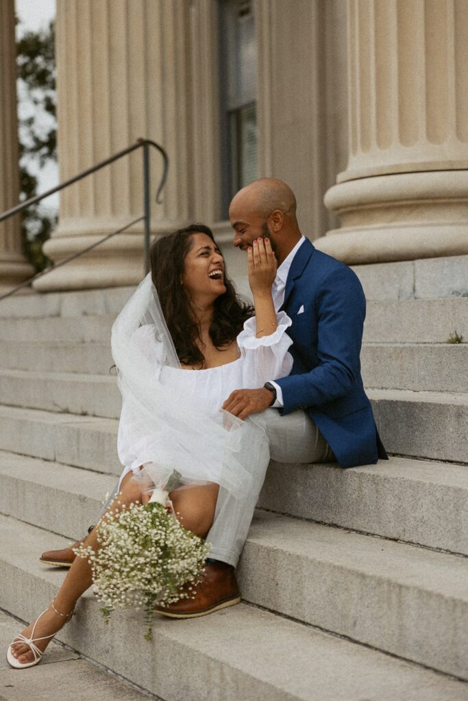 Man and woman sitting on courthouse steps and laughing in wedding attire. 