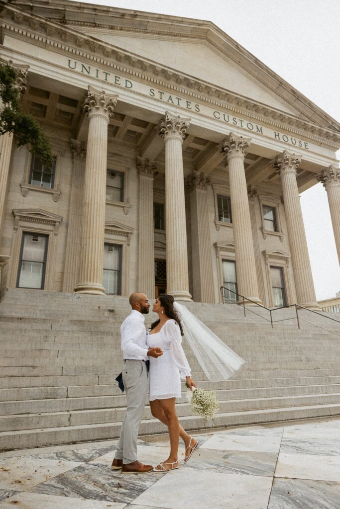Man and woman in wedding attire about to kiss in front of courthouse.