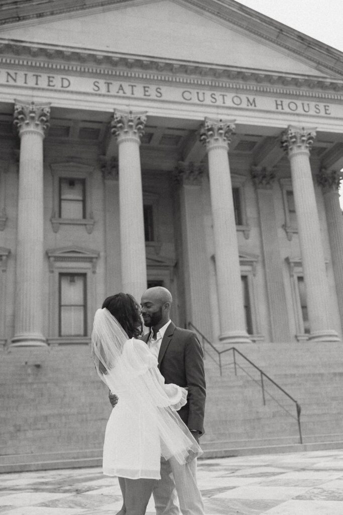 Man and woman in wedding attire with faces very close together in front of Charleston courthouse. 
