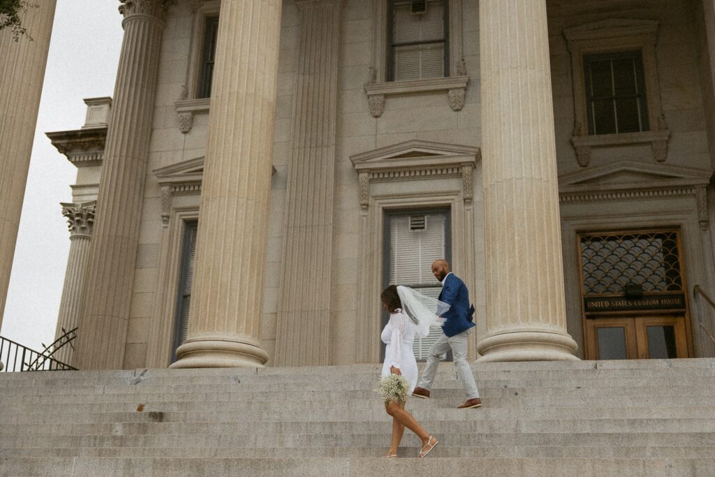 Man and woman walking across steps at courthouse while holding hands in wedding attire. 
