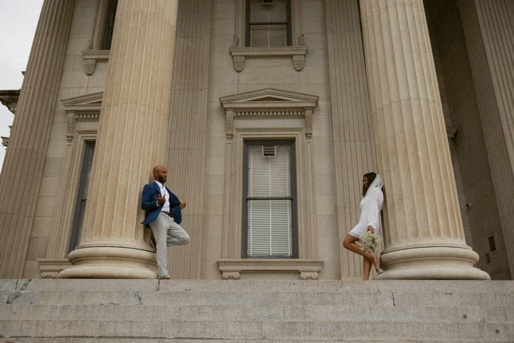Man and woman in wedding attire leaning on columns at courthouse looking at each other.