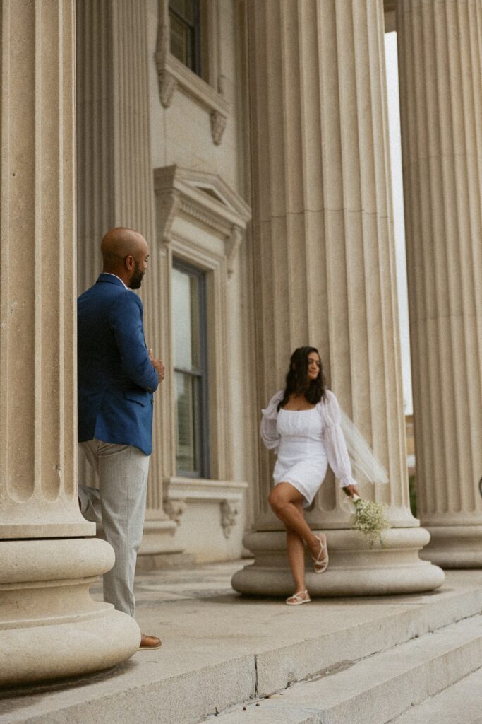 Man looking at woman in wedding dress while she is leaning on a column at courthouse.