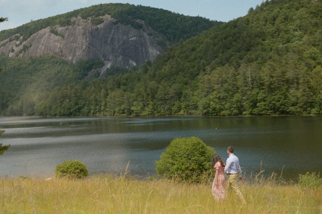 Man and woman walking through field in front of lake with mountains.
