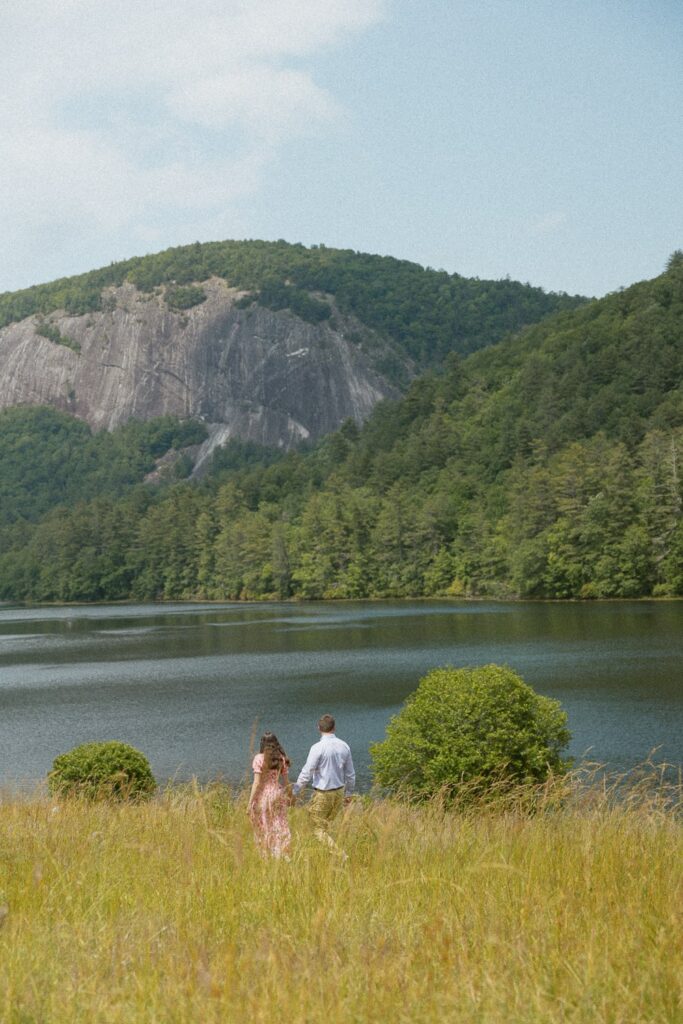 Man and woman walking through field in front of lake with mountains.