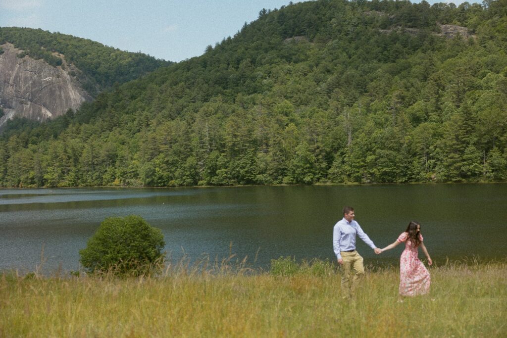 Woman pulling man by the hand through a field in front of a lake with mountains.