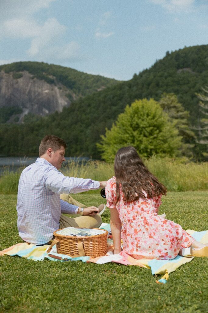 Man and woman sitting on blanket having a picnic in front of a lake with mountains.