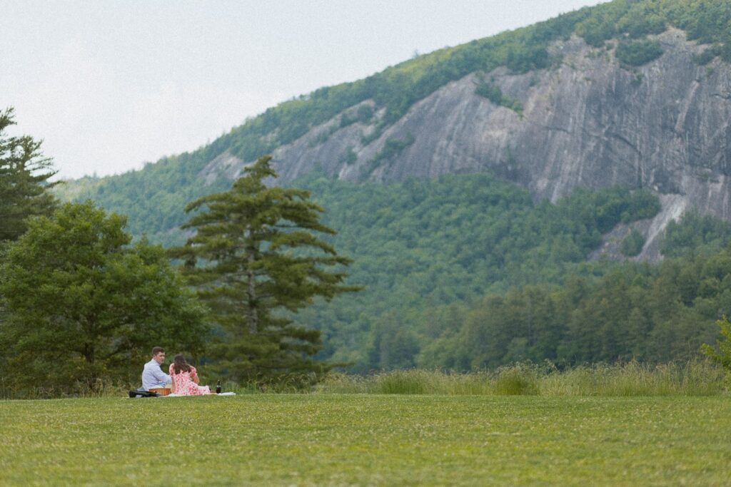 Man and woman sitting on blanket having a picnic in a field in front of mountain.