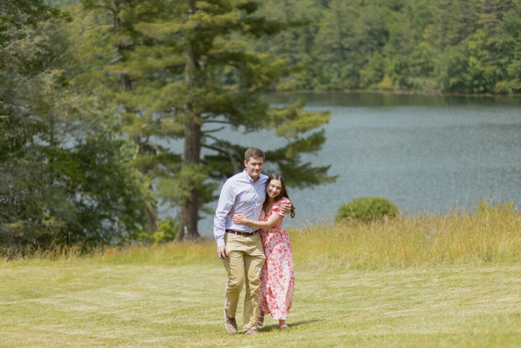 Man and woman hugging and walking through a field in front of a lake.