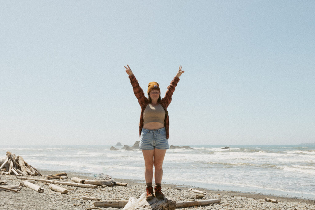 Woman standing on log in front of ocean holding up peace signs.