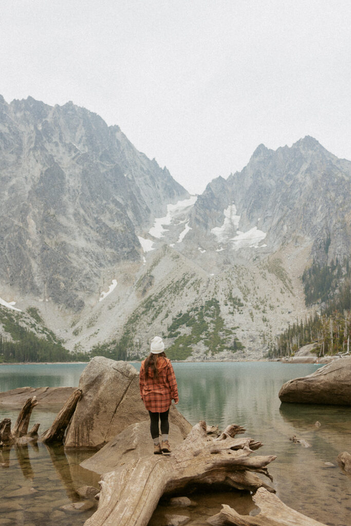 woman standing on log looking at alpine lake with snowcapped mountains.