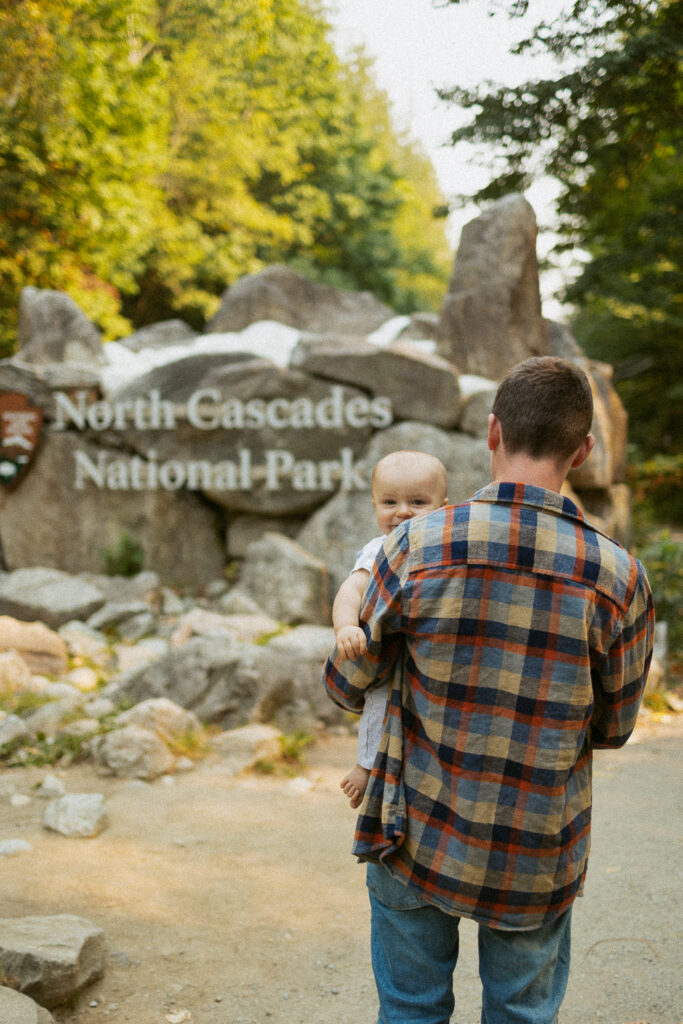 Boy holding a baby in front of north cascades national park entrance sign.