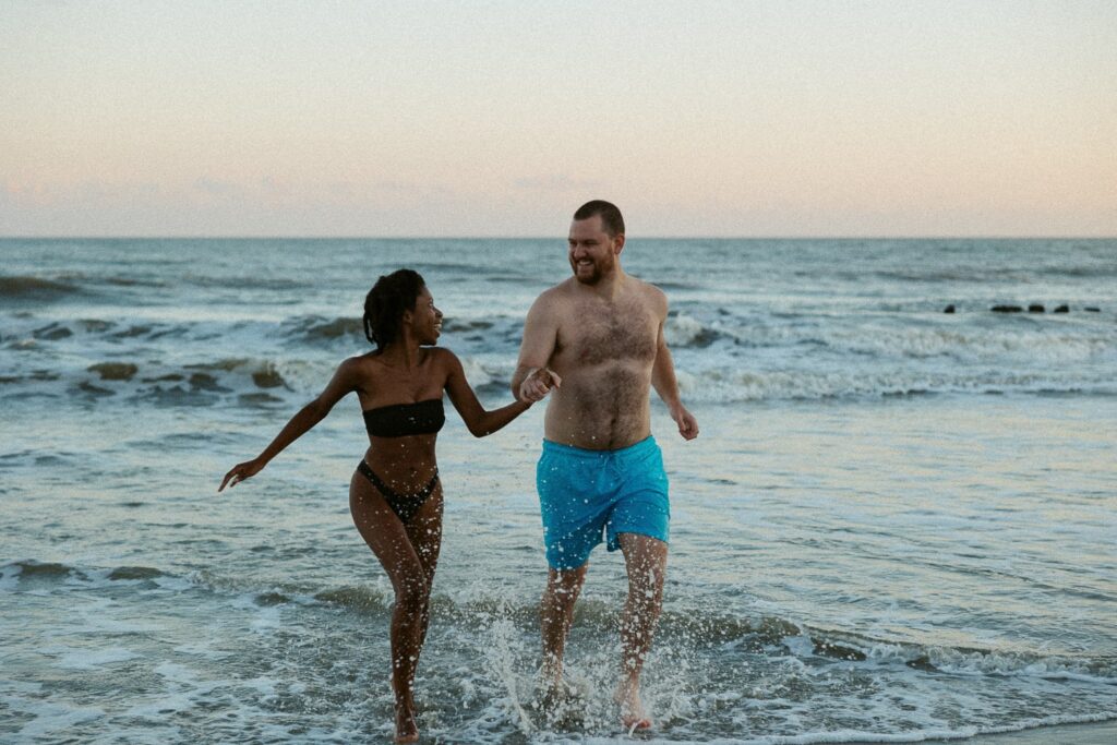 Man and woman holding hands and running in the ocean at sunset.