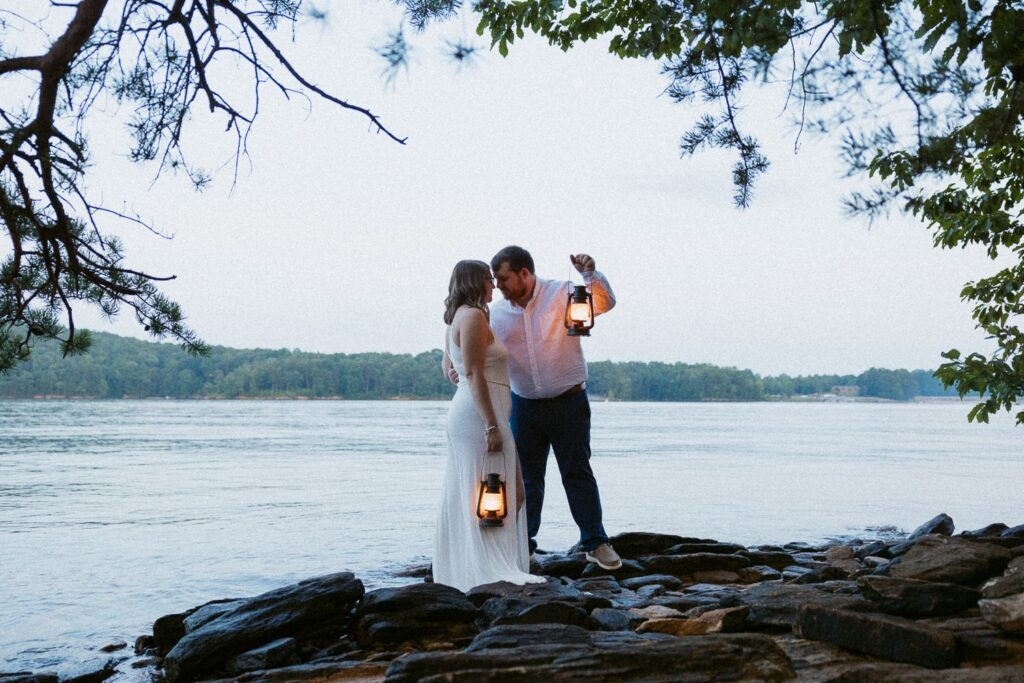 Man and woman standing on rocks in front of lake while holding lanterns.