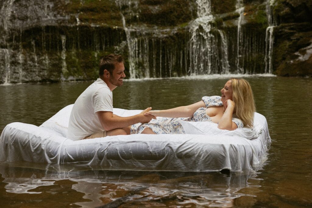 Man and woman on an air mattress in the water in front of a waterfall.