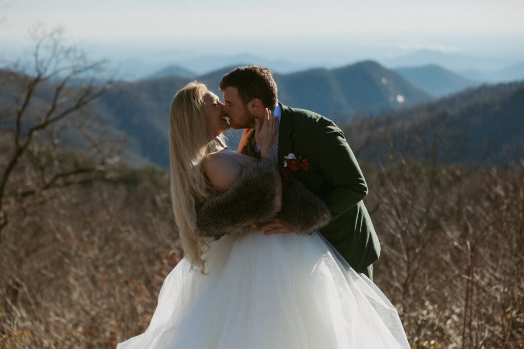 Man and woman about to kiss while dressed in wedding attire with mountains in the background. 