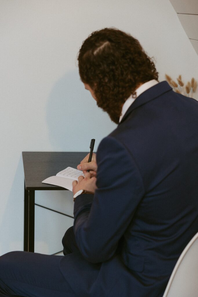 Man in wedding attire sitting and writing vows in a vow book.
