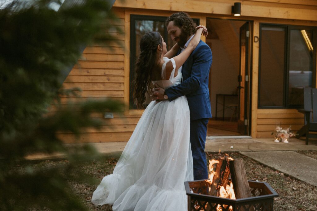 Man and woman in wedding attire dancing around campfire in front of airbnb. 