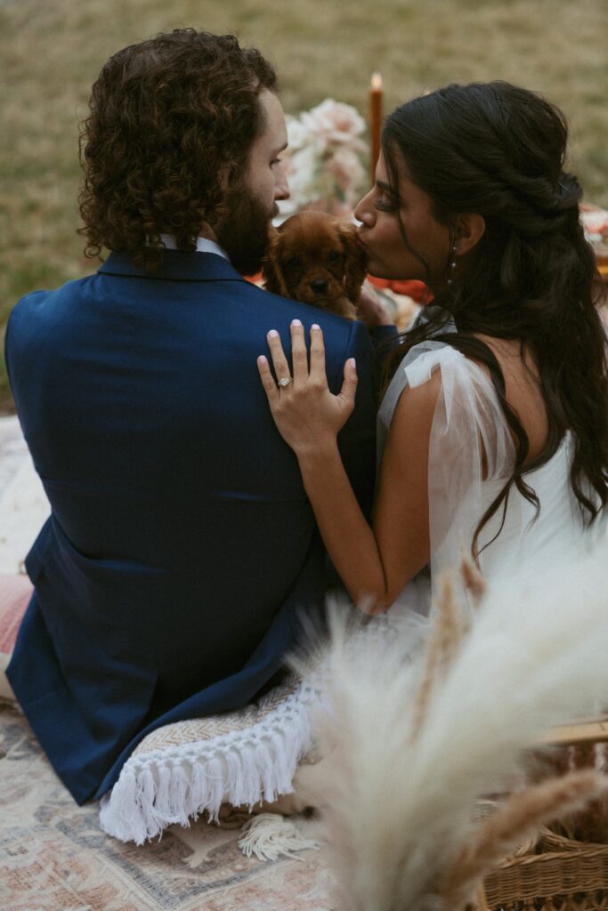 Man and woman kissing their dog during picnic at wedding reception.