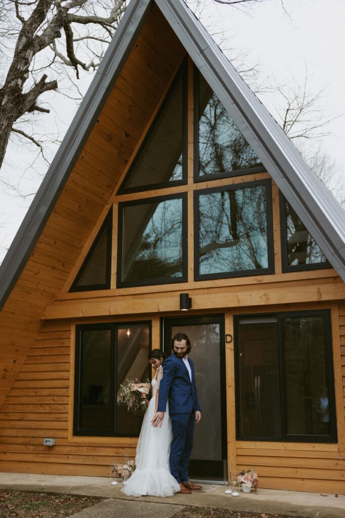 Man and woman in wedding attire standing back to back and holding hands in front of an a-frame cabin.