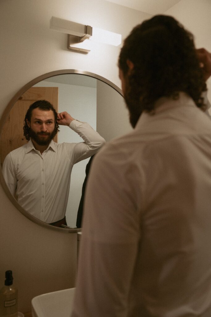 Man in wedding attire looking in a mirror and fixing hair. 
