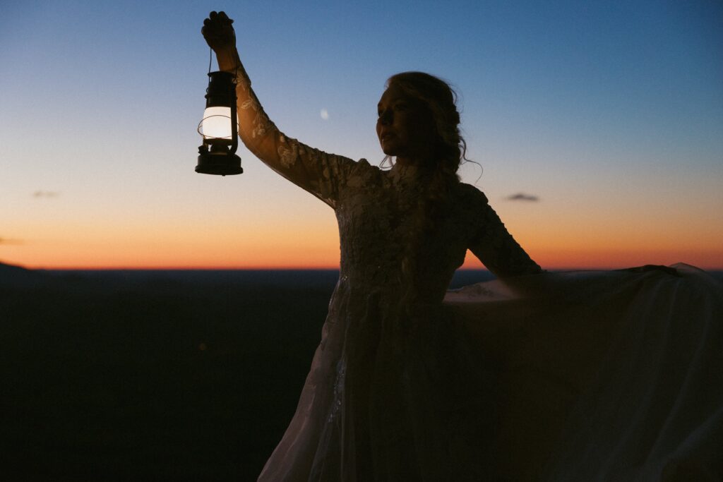 Woman in wedding dress holding lantern in front of moon and mountains at sunrise. 