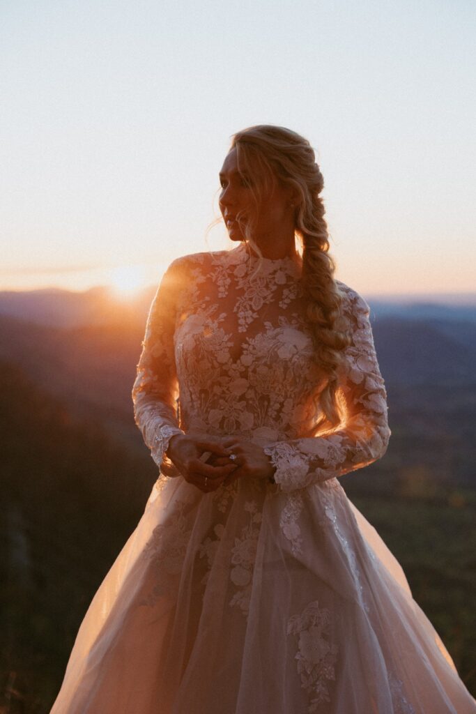 Woman in a wedding dress standing in front of mountains and sunrise while holding her engagement ring.