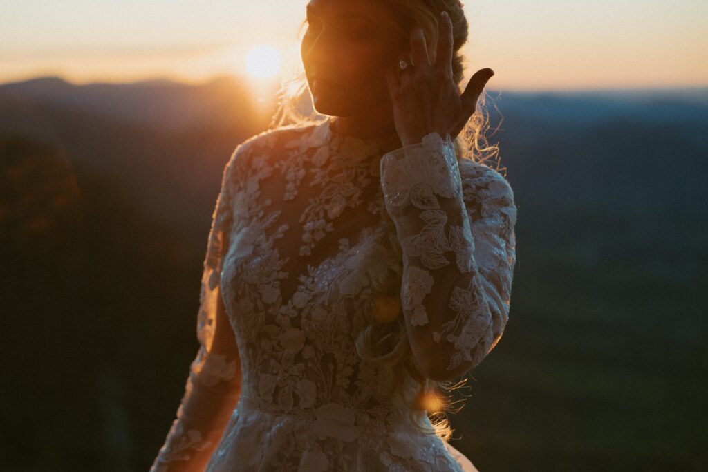 Woman in a wedding dress brushing her hair out of her face with the mountains and sunrise behind her. 