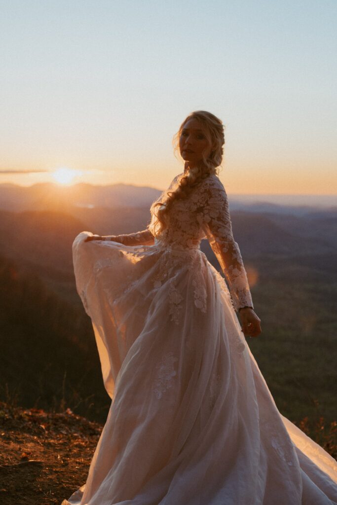Woman in a wedding dress spinning with mountains in the background while sun is coming over them. 